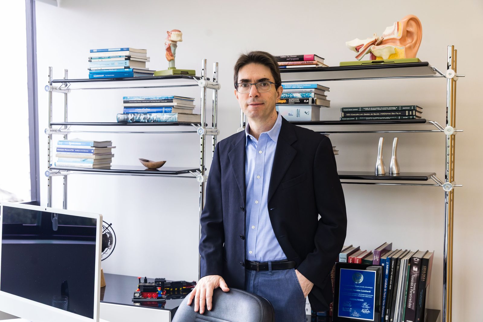 Dr. Fernando Danelon Leonhardt em seu consultório, ao lado de estante com livros e modelo anatômico de ouvido